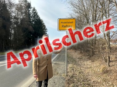 Auf dem Bild ist Michael Abraham vor dem Ortsschild "Modellstadt Rehau" zu sehen. Davor ist in rot geschrieben "Aprilscherz".