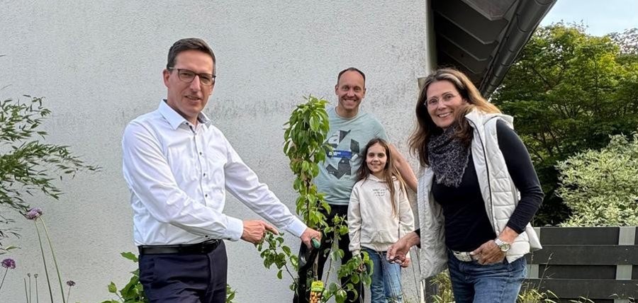 Auf dem Bild ist Bürgermeister Michael Abraham zu sehen, wie er einen Baum bei den Bauherren Kerstin Kropf und Markus Müller sowie deren Tochter Melissa in deren Garten pflanzt.