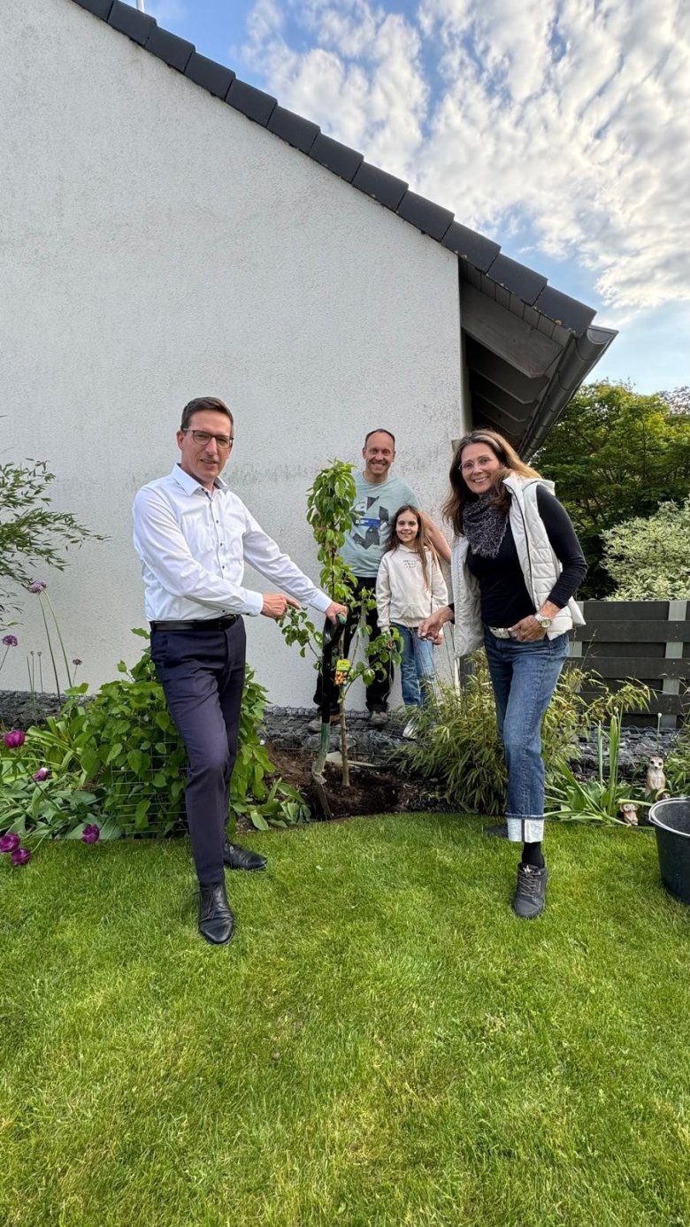 Auf dem Bild ist Bürgermeister Michael Abraham zu sehen, wie er einen Baum bei den Bauherren Kerstin Kropf und Markus Müller sowie deren Tochter Melissa in deren Garten pflanzt.