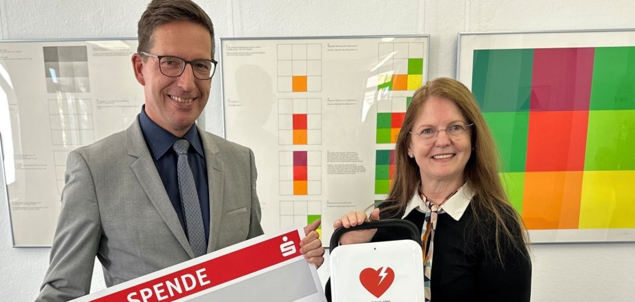 Das Bild zeigt Bürgermeister Abraham mit dem symbolischen Scheck neben 1. Vorsitzenden des Kunstvereins, Annie Sziegoleit, die den neuen Defibrillator in Händen hält.