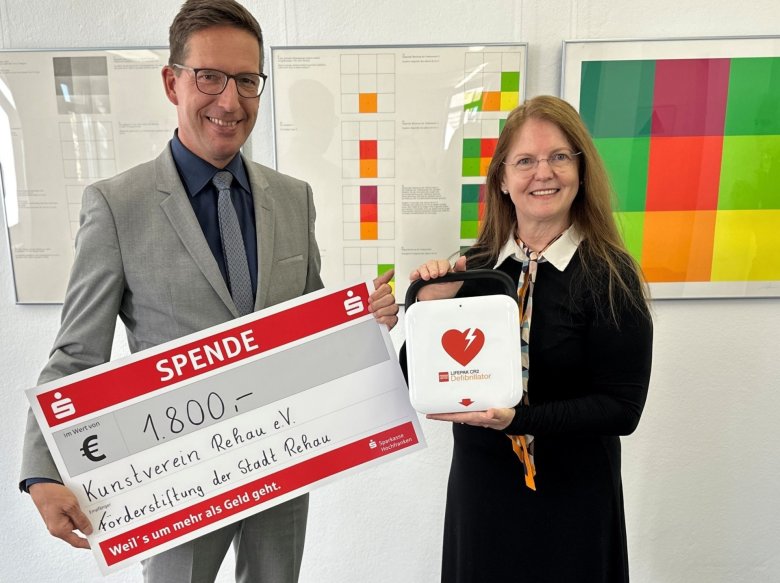 Das Bild zeigt Bürgermeister Abraham mit dem symbolischen Scheck neben 1. Vorsitzenden des Kunstvereins, Annie Sziegoleit, die den neuen Defibrillator in Händen hält.