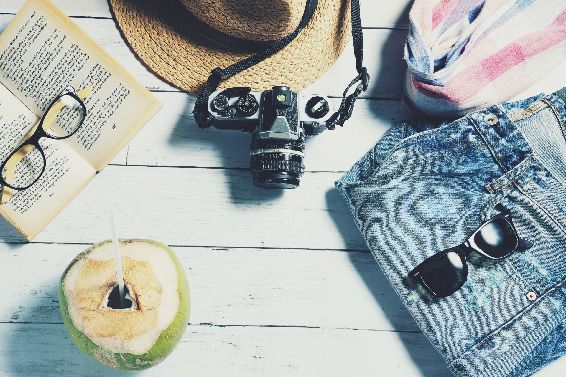 Auf dem Bild sind eine Sonnenbrille, eine Lesebrille, eine Kokosnuss, ein Sonnenhut, eine Jeans und eine Kamera zu sehen.