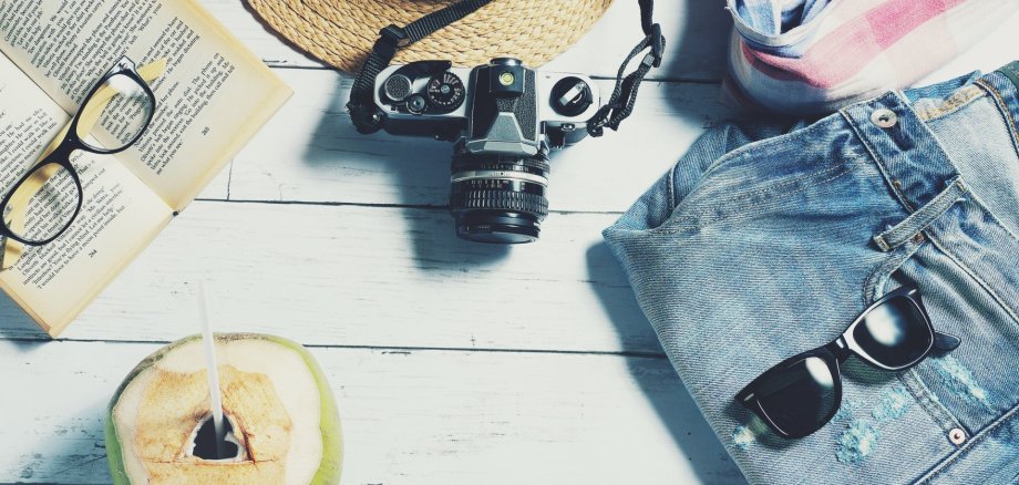 Auf dem Bild sind eine Sonnenbrille, eine Lesebrille, eine Kokosnuss, ein Sonnenhut, eine Jeans und eine Kamera zu sehen.