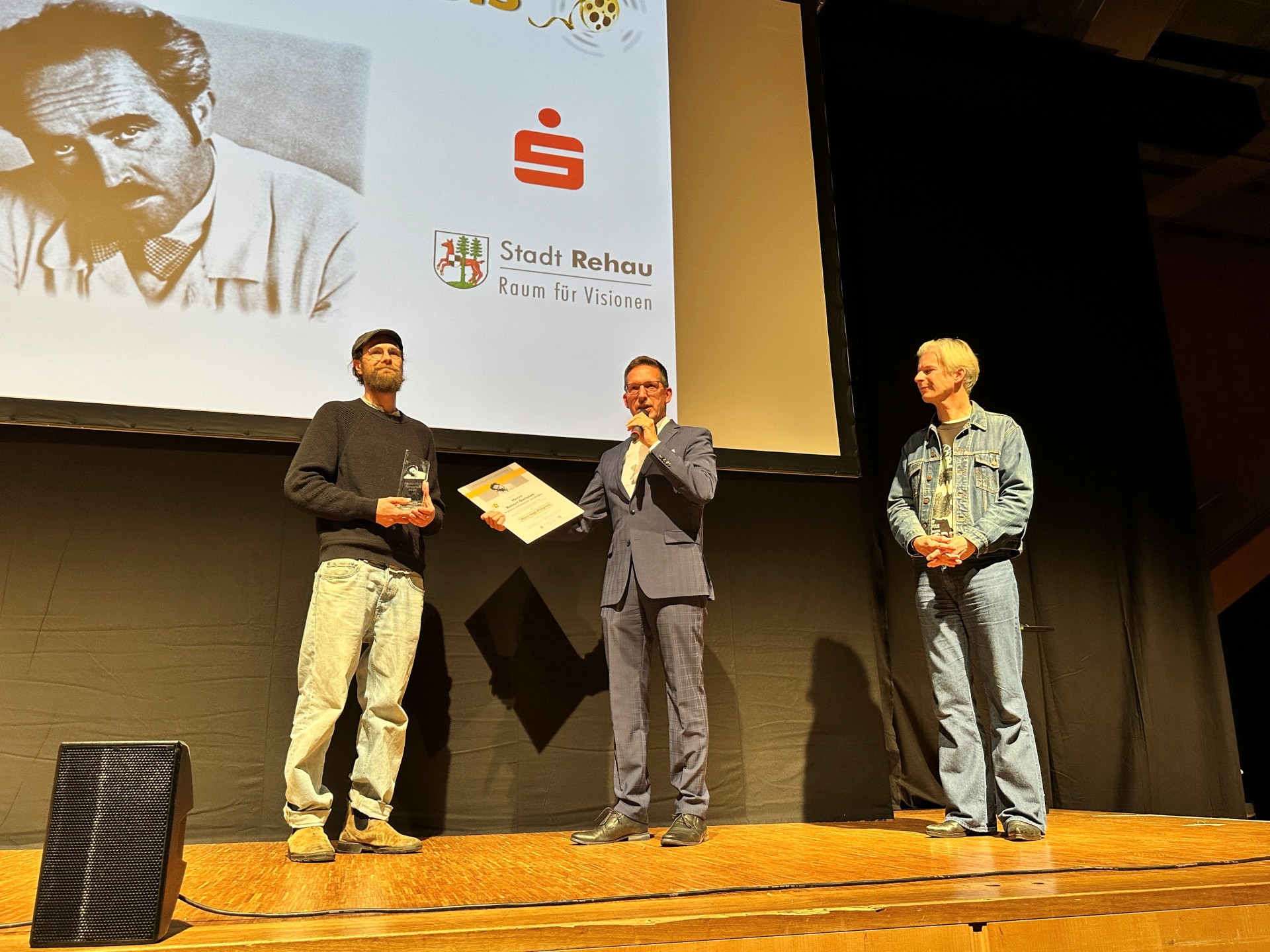 Auf dem Bild sind Bürgermeister Michael Abraham und Thorsten Schaumann neben dem diesjährigen Hans-Vogt-Filmpreisträger Robert Gwisdek zu sehen, der gerade die Urkunde und den Preis von Bürgermeister Abraham auf der Bühne entgegennimmt.