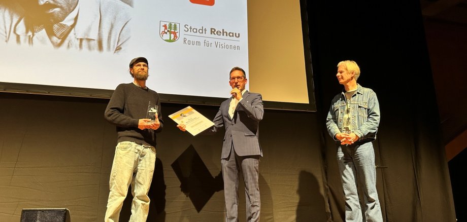 Auf dem Bild sind Bürgermeister Michael Abraham und Thorsten Schaumann neben dem diesjährigen Hans-Vogt-Filmpreisträger Robert Gwisdek zu sehen, der gerade die Urkunde und den Preis von Bürgermeister Abraham auf der Bühne entgegennimmt.