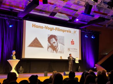 Auf dem Bild ist Erster Bürgermeister Michael Abraham auf der Bühne der Freiheitshalle zu sehen, wie er ein Palakt hochhält und zum Publikum spricht. an der Seite wartet Thorsten Schaumann von den Hofer Filmtagen. Im Hintergrund ist eine Leinwand zu sehen, mit einer Grafik auf der "Hans-Vogt-Filmpreis" steht.