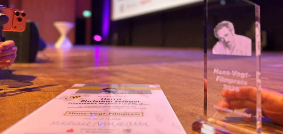Auf dem Bild ist der Hans-Vogt-Filmpreis sowie die zugehörige Urkunde zu sehen. Der Name des diesjährigen Preisträgers ist auf beiden zu sehen: Christian Friedel.