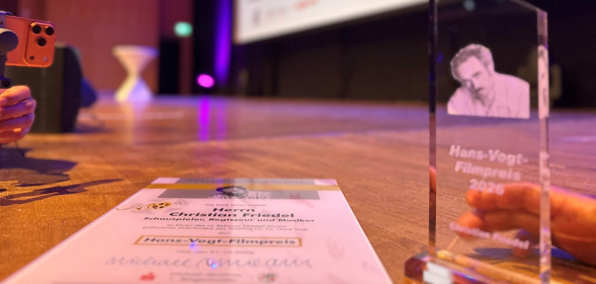 Auf dem Bild ist der Hans-Vogt-Filmpreis sowie die zugehörige Urkunde zu sehen. Der Name des diesjährigen Preisträgers ist auf beiden zu sehen: Christian Friedel.