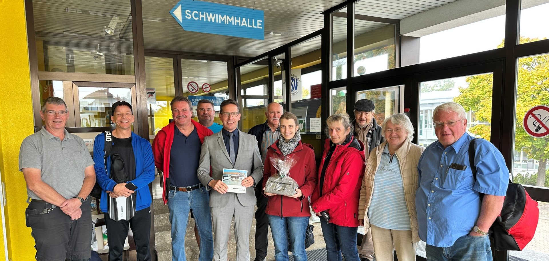 Das Bild zeigt Julia Nüßel, die erste Besucherin des Hallenbads 2025/2026 neben weiteren Badegästen, der Badeaufsicht, dem Hausmeister Wolfgang Meichner sowie 1. Bürgermeister Michael Abraham.