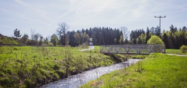 Das Bild zeigt eine Brücke der Perlenroute und dem neu geschaffenen Themenwanderwegs über einen Bach, Wiese links und rechts sowie die Bahnschiene links.