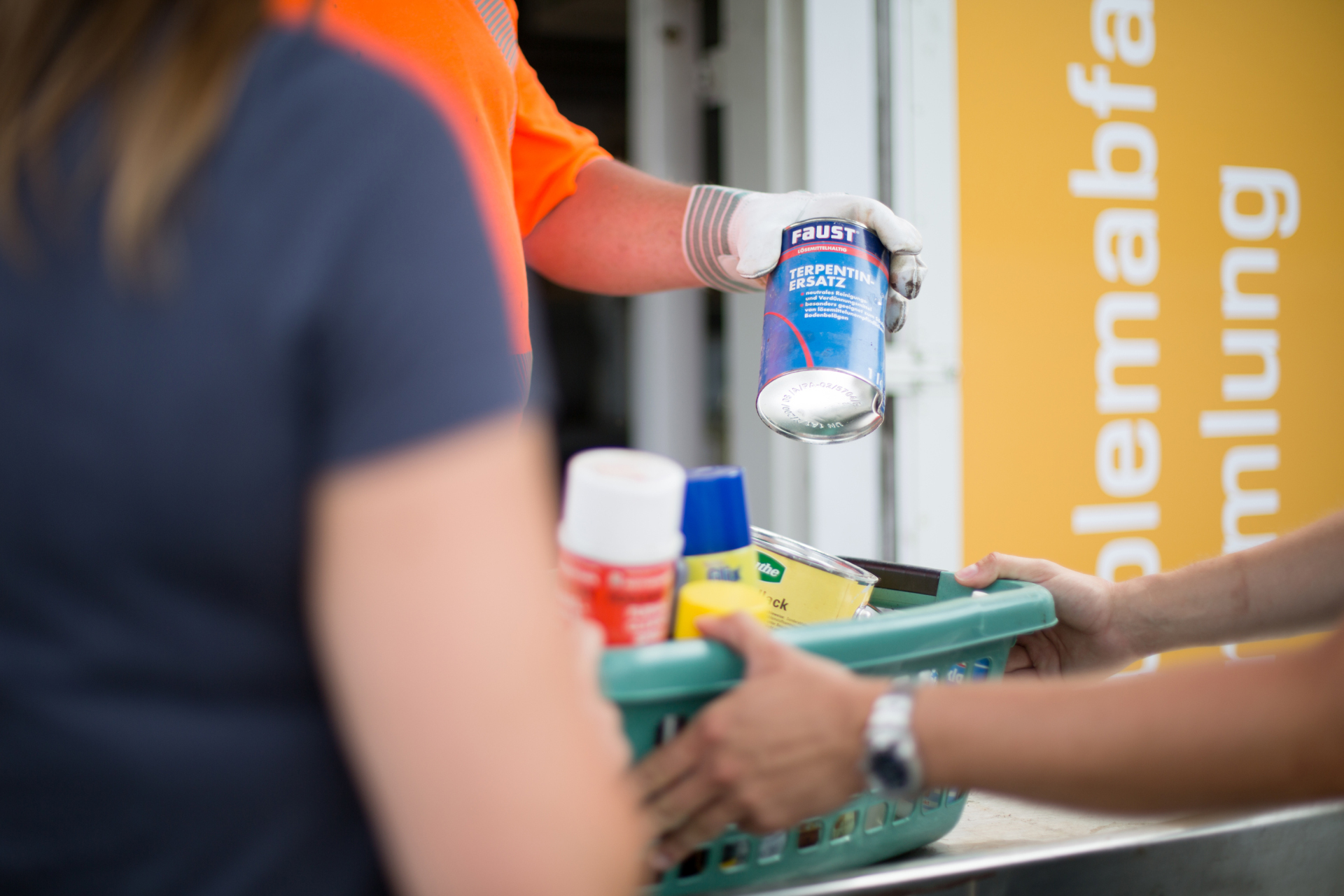 Das Bild zeigt mittig einen Plastik-Korb, in welchem sich verschiedene Dosen und Sprühflaschen befinden. Eine Person hält diesen Korb, eine entnimmt gerade eine Dose. Im Hintergrund ist auf der gelben Fassade das Wort "Problemabfallsammlung" zu erkennen.