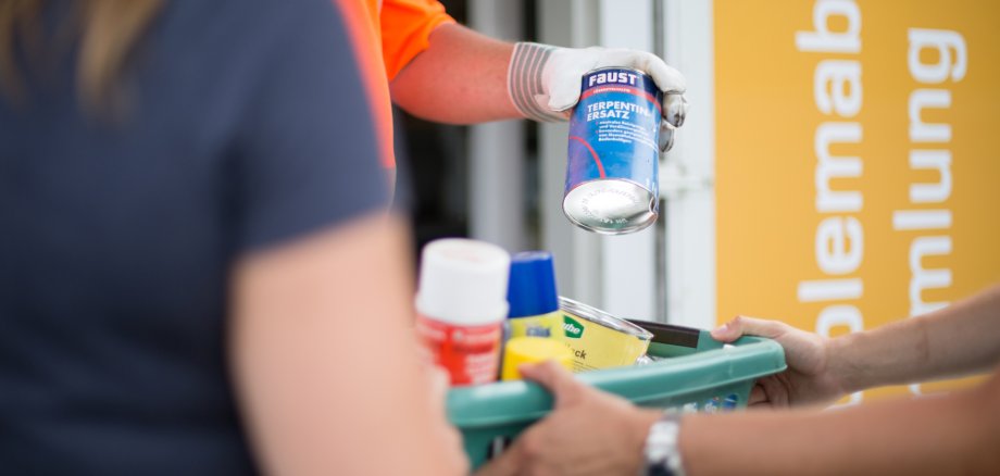 Das Bild zeigt mittig einen Plastik-Korb, in welchem sich verschiedene Dosen und Sprühflaschen befinden. Eine Person hält diesen Korb, eine entnimmt gerade eine Dose. Im Hintergrund ist auf der gelben Fassade das Wort "Problemabfallsammlung" zu erkennen.