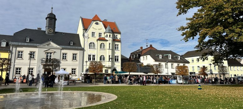 Auf dem Bild ist im Vordergrund die Wiese auf dem Maxplatz in Rehau zu sehen. Dahinter sind einige Marktstände sowie das Alte Rathaus Rehau zu erkennen. Auf der Wiese befindet sich etwas Laub, das auf die Jahreszeit Herbst hindeutet.