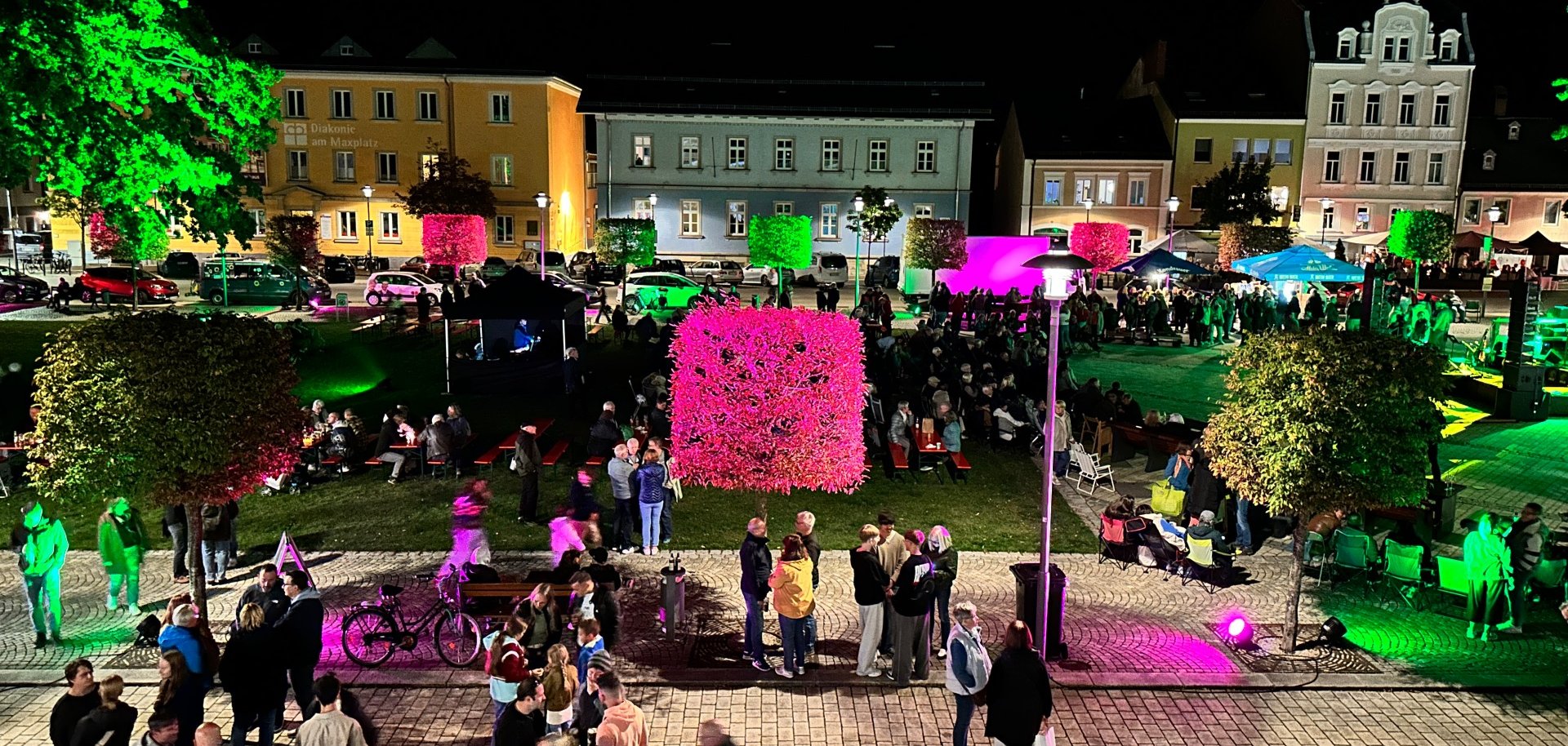 Das Bild zeigt den belebten Maxplatz bei Nacht, Blickwinkel vom Festsaal des Alten Rathauses aus. Die Bäume sind farbig beleuchtet.