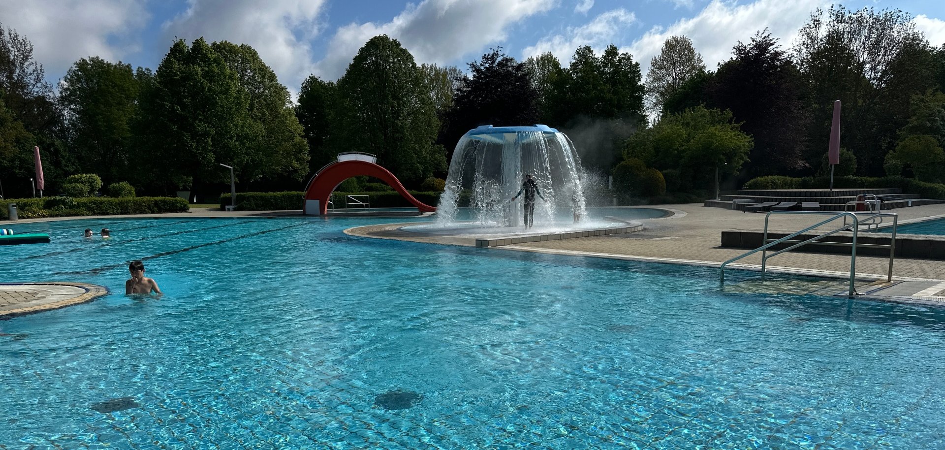 Das Bild zeigt das Nichtschwimmerbecken im Rehauer Freibad. Im Hintergrund ist der "Pilz" zu sehen, der gerade Wasser spritzt und ein Kind sich darunter befindet.