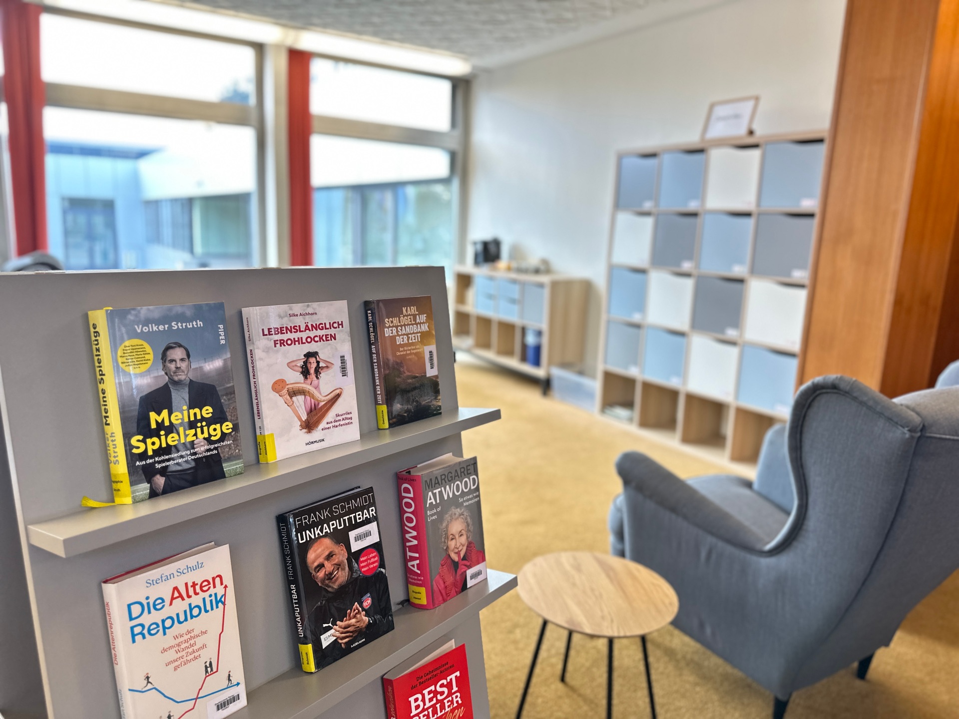 Das Bild zeigt im Vordergrund Bücher auf einem grauen Aufsteller, daneben ist ein bequemer Sessel sowie im Hintergrund eine Kaffee-Ecke zu erkennen.