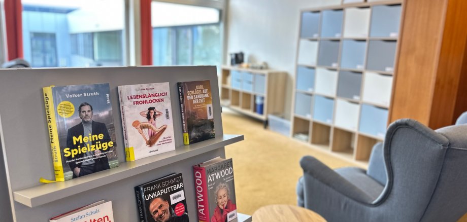 Das Bild zeigt im Vordergrund Bücher auf einem grauen Aufsteller, daneben ist ein bequemer Sessel sowie im Hintergrund eine Kaffee-Ecke zu erkennen.