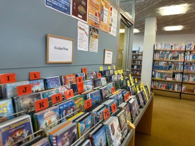 Auf dem Bild sind DVDs, geordnet nach Anfangsbuchstabe zu sehen. Darüber ist ein Schild mit der Aufschrift "Kinderfilme" platziert. Im Hintergrund sind weitere Filme und viele Bücher in Regalen zu sehen.