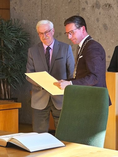 Auf dem Bild sind 2. Bürgermeister Rudolf Scholz und Bürgermeister Michael Abraham zu sehen. Abraham überreicht Herrn Scholz eine DinA3-Urkunde.