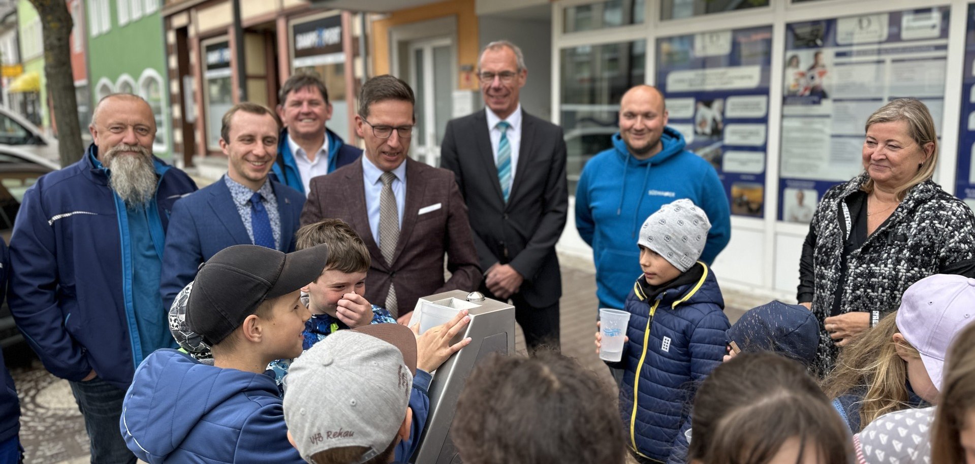 Das Bild zeigt den neuen Trinkwasserbrunnen mittig, während der Becher eines Kindes mit Wasser gefüllt wird. Außerdem sind weitere Kinder des Kinderhorts des MGH vor Ort sowie Bürgermeister Michael Abraham, Werkleiter Beckstein, Kämmerer Muggenthaler und Mitarbeiter der Firma SüdWasser.