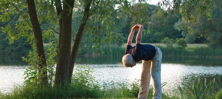 Das Bild zeigt einen älteren Herren auf einer Wiese vor einem See. Er dehnt sich gerade nach unten und hält seine Hände zurückgefalten nach oben.