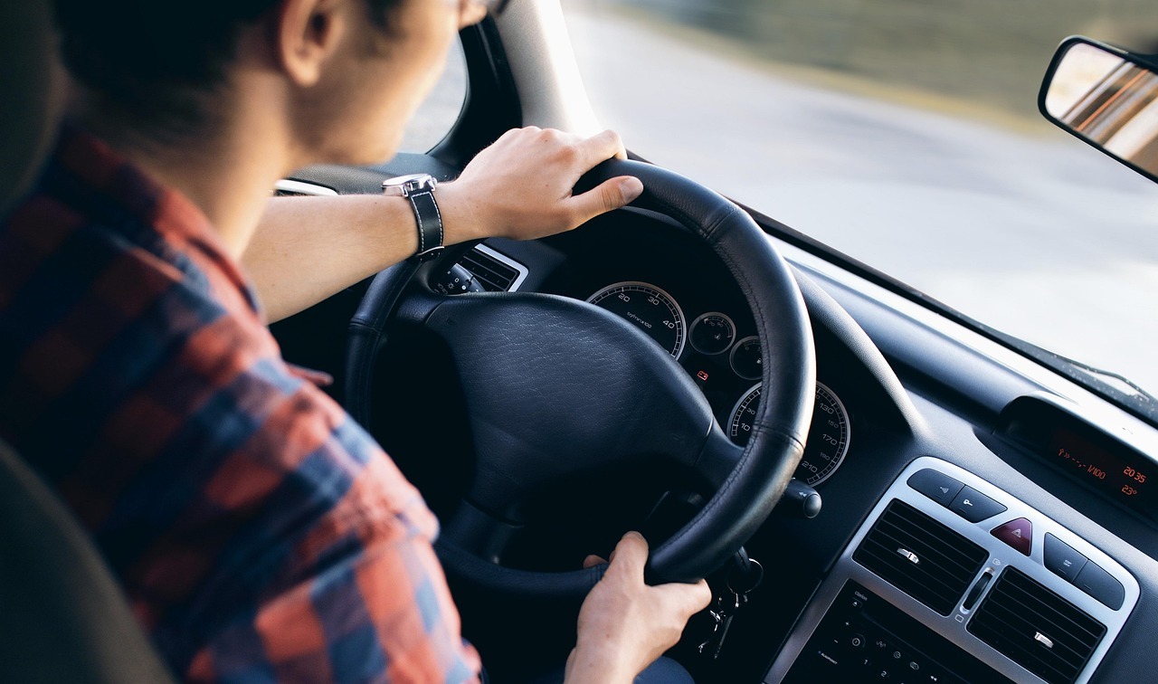 Das Bild zeigt einen Herren am Steuer eines Autos, während er gerade das Lenkrad mit beiden Händen umfasst.