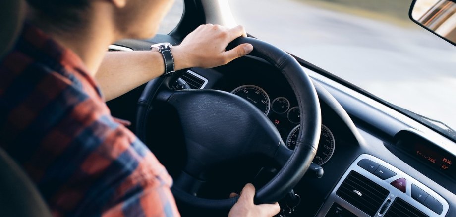 Das Bild zeigt einen Herren am Steuer eines Autos, während er gerade das Lenkrad mit beiden Händen umfasst.