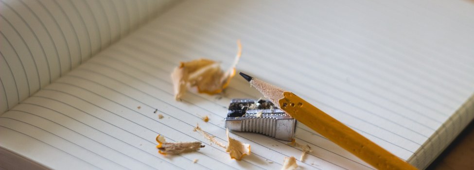 Das Bild zeigt einen gespitzten Bleistift auf einem Metall-Spitzer. Daneben befinden sich die Späne, also der Abfall vom Spitzen und darunter ein liniertes Notizbuch.