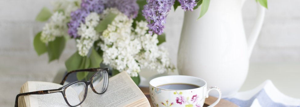 Das Bild zeigt einen runden Holzteller, auf welchem sich eine kleine Kaffeetasse und ein aufgeschlagenes Buch sowie eine Lesebrille befindet. Im Hintergrund sind weiße und lilafarbige Blumen zu sehen.