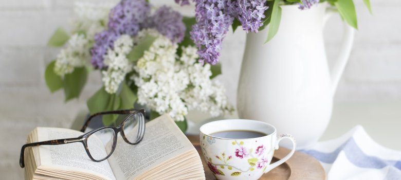 Das Bild zeigt einen runden Holzteller, auf welchem sich eine kleine Kaffeetasse und ein aufgeschlagenes Buch sowie eine Lesebrille befindet. Im Hintergrund sind weiße und lilafarbige Blumen zu sehen.