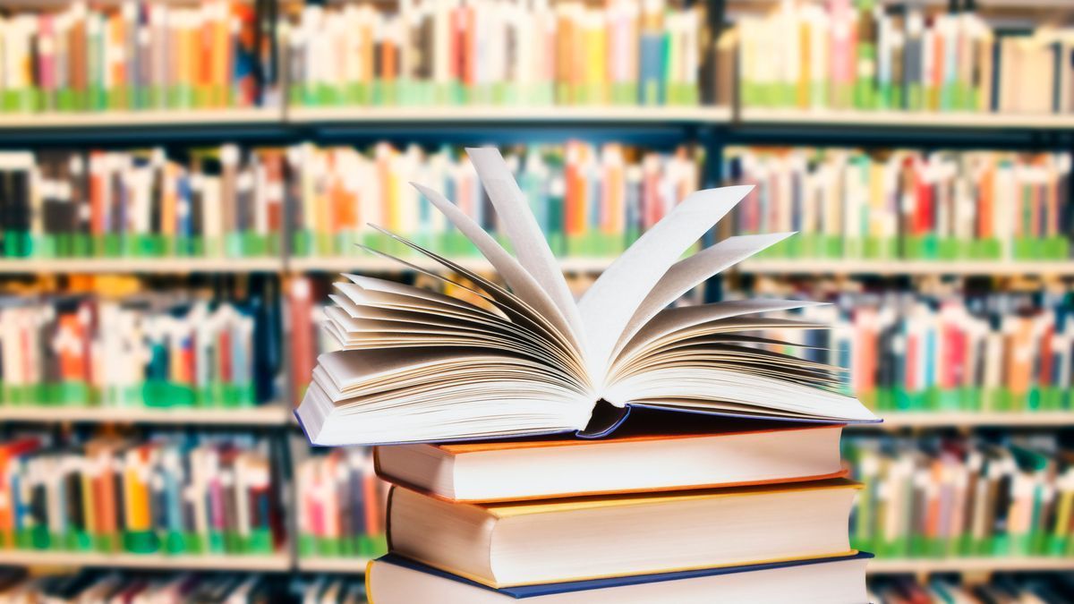 Das Bild zeigt ein aufgeschlagenes Buch, welches auf einem Bücherstapel liegt. Im Hintergrund ist ein Regal mit vielen Büchern zu erkennen, welche alle mit einem grünen Aufkleber am Band versehen sind - es handelt sich vermutlich um eine Bücherei.