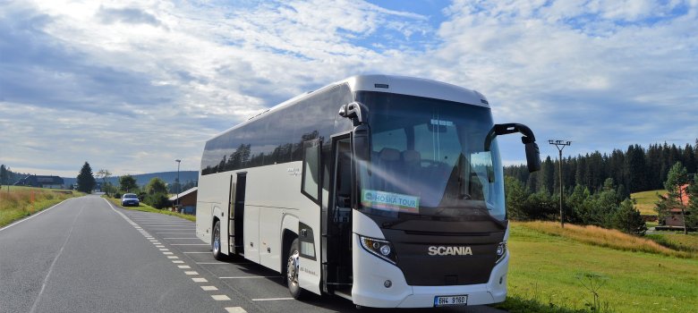 Das Bild zeigt einen weißen Reisebus, welcher seine Türen gerade geöffnet hat und neben einer Straße platziert ist. Rechts ist eine Wiese zu erkennen, im Hintergrund auch einige wenige Anwesen.