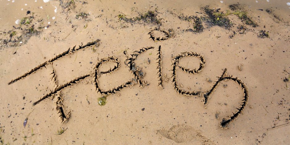 Das Bild zeigt einen Strand mit Sand, hintergründig sind ein wenig Algen zu erkennen. Im Sand ist das Wort "Ferien" zu erkennen.