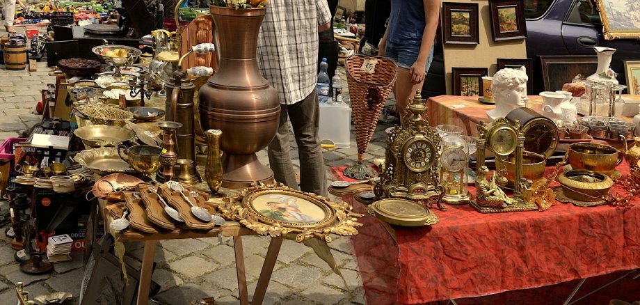 Das Bild zeigt mehrere Flohmarkt-Stände mit Trödel-Artikeln wie alte Standuhren, Silberlöffel oder Bilder mit hochwertigen Rahmen.