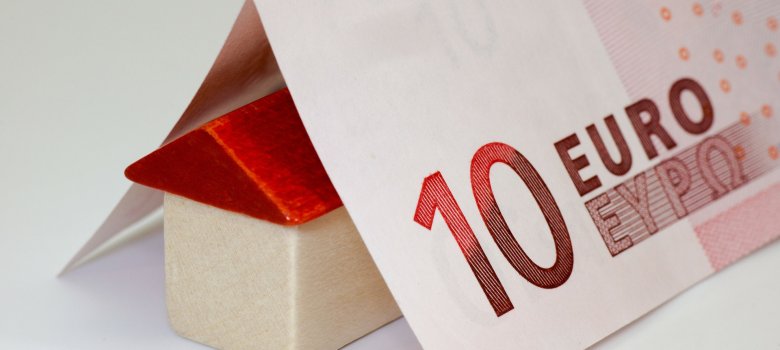 Das Bild zeigt einen gefaltenen 10-Euro-Schein, welcher auf einem Holzwürfel und -dreieck, welche ein Haus darstellen, liegt.