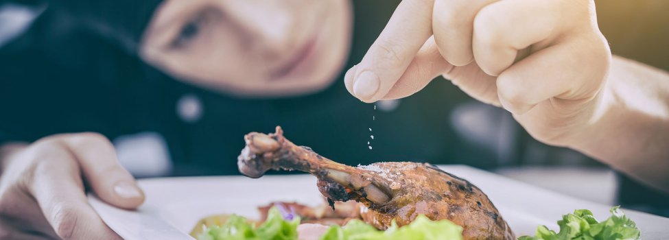 Das Bild zeigt im Vordergrund einen eckigen Teller mit Salatblättern, darauf ein knuspriger Hähnchenschenkel, welcher gerade von der Köchin, die im Hintergrund zu sehen ist, mit etwas bestreut wird.