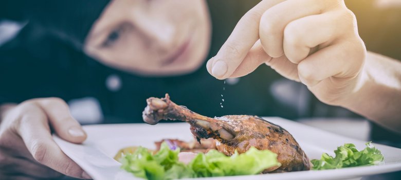 Das Bild zeigt im Vordergrund einen eckigen Teller mit Salatblättern, darauf ein knuspriger Hähnchenschenkel, welcher gerade von der Köchin, die im Hintergrund zu sehen ist, mit etwas bestreut wird.