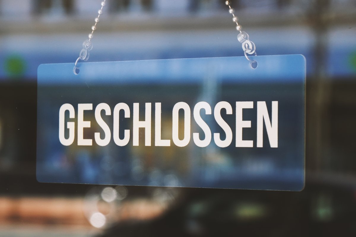 Das Bild zeigt ein Glas-Schild, auf welchem "GESCHLOSSEN" steht und an einer Kette von oben befestigt ist.