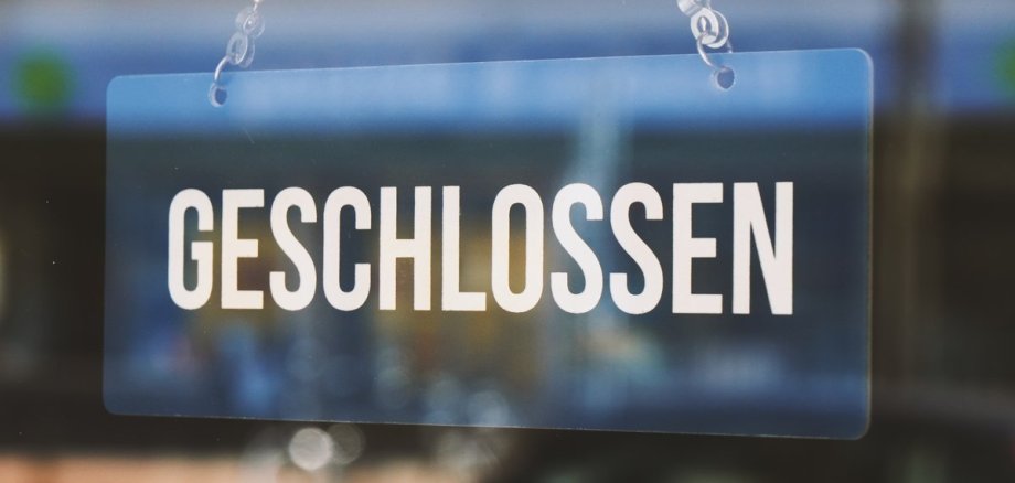 Das Bild zeigt ein Glas-Schild, auf welchem "GESCHLOSSEN" steht und an einer Kette von oben befestigt ist.