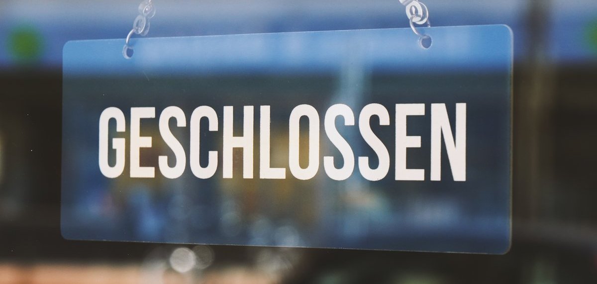 Das Bild zeigt ein Glas-Schild, auf welchem "GESCHLOSSEN" steht und an einer Kette von oben befestigt ist.