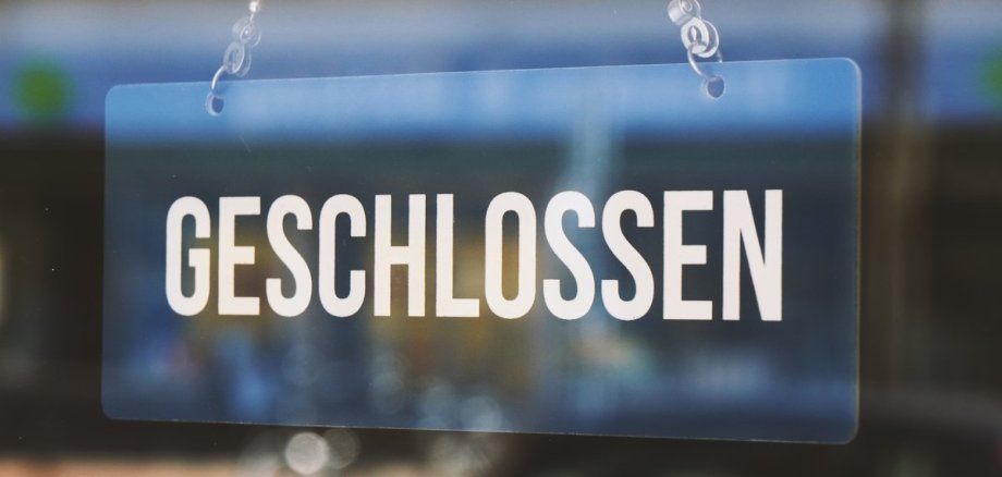Das Bild zeigt ein Glas-Schild, auf welchem "GESCHLOSSEN" steht und an einer Kette von oben befestigt ist.