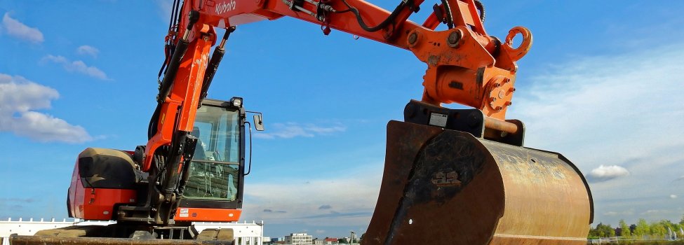 Auf dem Bild ist vordergründig ein orangefarbener Bagger zu sehen, welcher offensichtlich schon etwas gegraben hat. Außerdem ist ein bereits ausgeschaufelter schmaler, gerader Schacht in der Erde zu erkennen. Im Hintergrund sind Gebäude zu sehen.