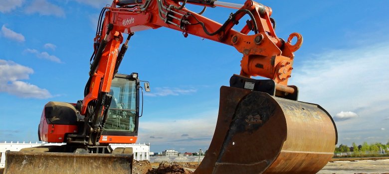 Auf dem Bild ist vordergründig ein orangefarbener Bagger zu sehen, welcher offensichtlich schon etwas gegraben hat. Außerdem ist ein bereits ausgeschaufelter schmaler, gerader Schacht in der Erde zu erkennen. Im Hintergrund sind Gebäude zu sehen.