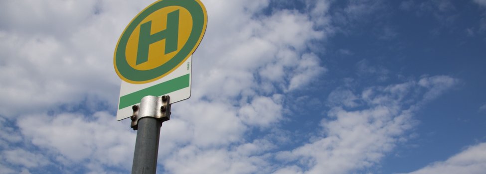 Das Bild zeigt ein grün-gelbes Haltestellenschild. Das Bild wurde von weiter unten aufgenommen und zeigt im Hintergrund den blauen Himmel mit einigen Wolken.