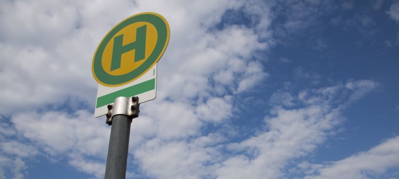 Das Bild zeigt ein grün-gelbes Haltestellenschild. Das Bild wurde von weiter unten aufgenommen und zeigt im Hintergrund den blauen Himmel mit einigen Wolken.