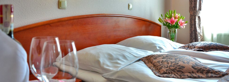 Das Bild zeigt ein Hotelzimmer. Zu sehen sind im Vordergrund zwei Weingläser sowie eine Flasche Wein. Dahinter befindet sich ein ordentlich gemachtes Bett und auf dem Nachtisch dahinter schöne rosafarbige Blumen.