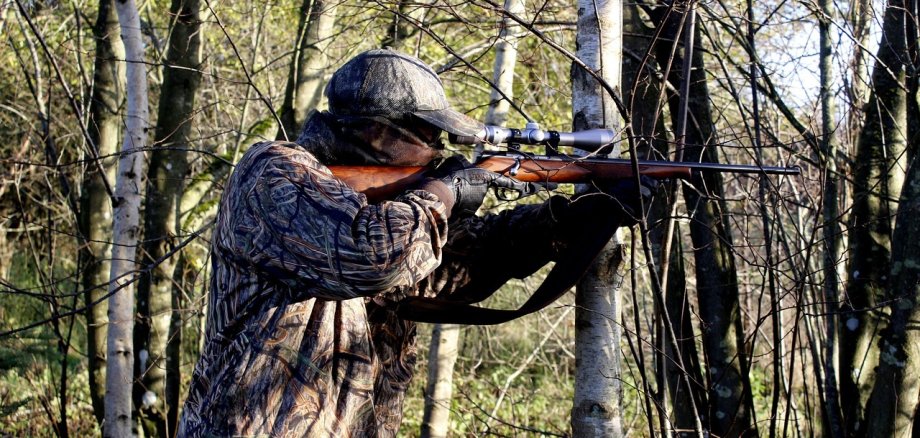 Das Bild zeigt eine Person, welche dank ihrer Klamotten inklusive der Cappie im Wald gut getarnt erscheint. Sie hat gerade ein Gewehr ausgerichtet.