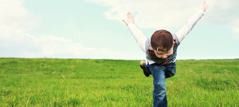 Das Bild zeigt ein Kind, welches gerade fröhlich über eine Wiese läuft, den Oberkörper nach vorne gebeugt hat und die Arme dabei nach hinten, bzw. oben streckt.