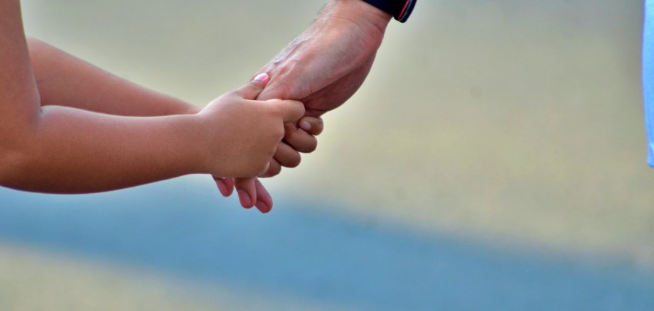Das Bild zeigt links zwei Arme eines Kindes, von welchen die Händen eine andere Hand eines Erwachsenen greifen, welcher sich rechts am Bild befindet.
