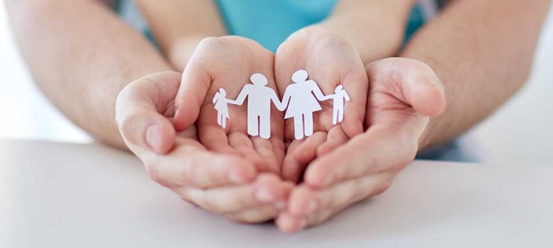 Das Bild zeigt vier Hände: Unten liegen die größeren Hände, vermutlich eines Mannes, nebeneinander mit den Handinnenflächen nach oben. Darüber sind ebenso zwei Hände nebeneinander gelegt. In den oberen Handinnenflächen befindet sich ein Papierzuschnitt, der eine Familie darstellen soll.